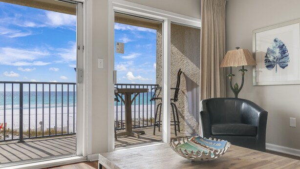 A room with a wooden table, a black leather chair, a beach view through a glass door, a framed leaf artwork, and a lamp. - Windancer 211. Gulf Front, Free Beach Service, Emerald Waters, Pool, Grill. (Miramar Beach)