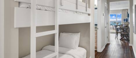 A bunk bed room with white bedding, a wooden floor, and a view of the balcony.