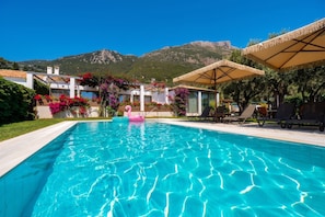 Ein Swimmingpool mit klarem, blauem Wasser, umgeben von Liegestühlen und Sonnenschirmen, mit Bergen im Hintergrund.