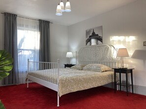 A bedroom with a white metal bed frame, a red carpet, a large window with sheer curtains, a nightstand with a lamp, and a wall-mounted picture of the Eiffel Tower. - Beautiful Second floor Furnished apartment in the heart of Newmarket (Newmarket)