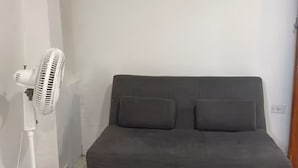 Une pièce avec un canapé gris et un ventilateur sur pied.