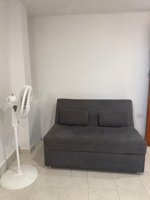 Een kamer met een grijze bank en een staande ventilator.