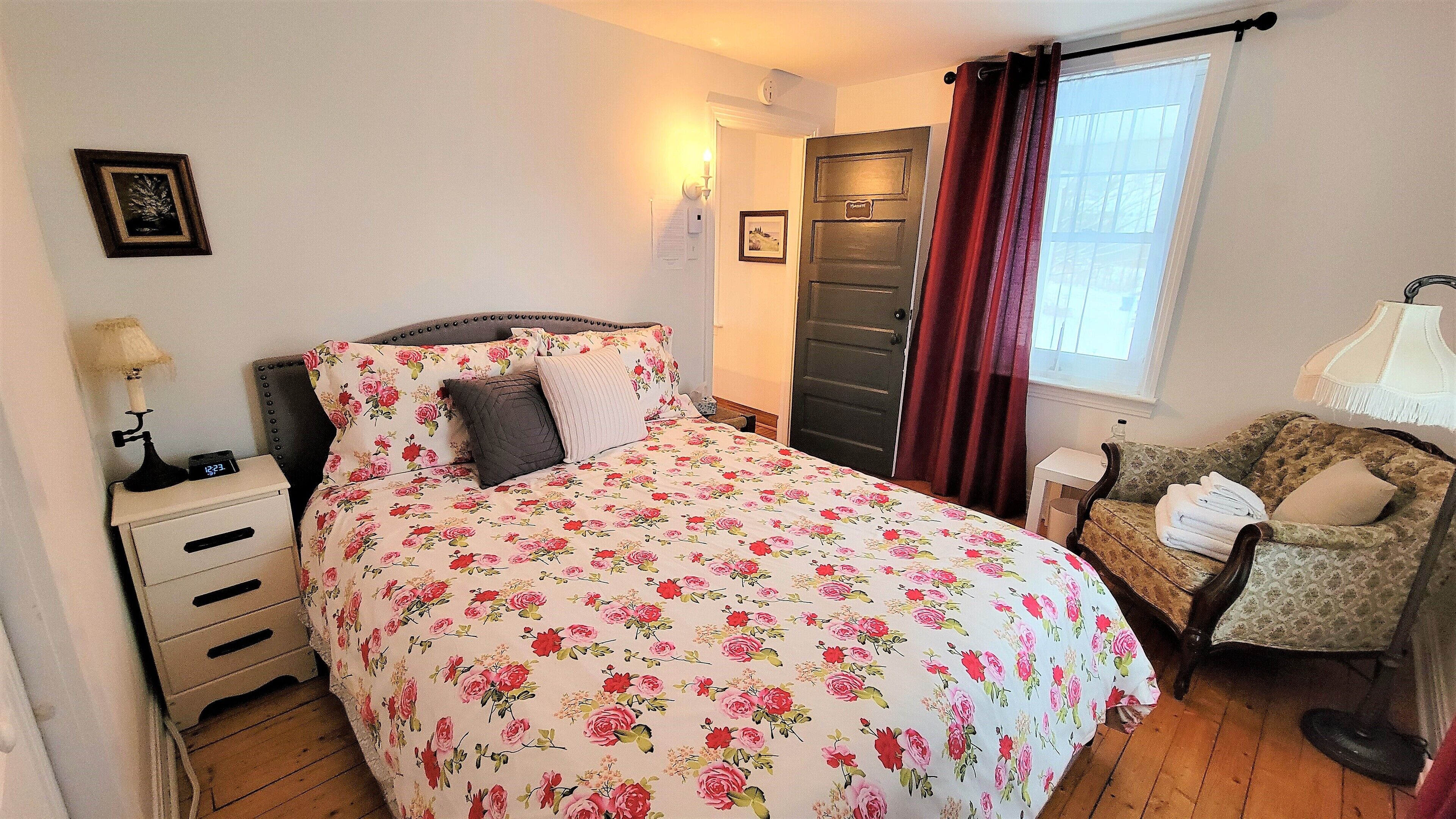 Un dormitorio con colcha estampada, una mesita de noche con lámpara, una silla con cojín y una puerta con manija.