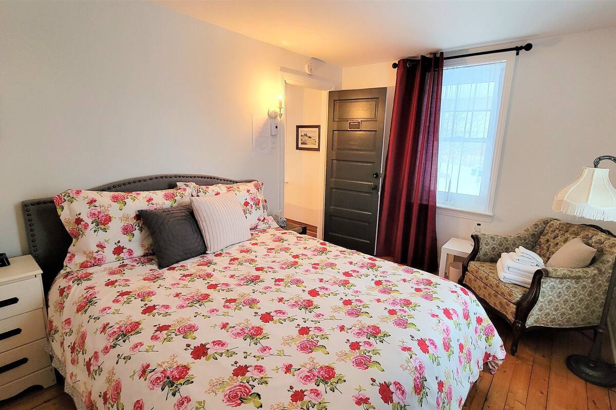 Une chambre à coucher avec une couvre-lit à motifs floraux, une table de chevet avec une lampe, une chaise avec des serviettes et une porte avec une plaque nominative.