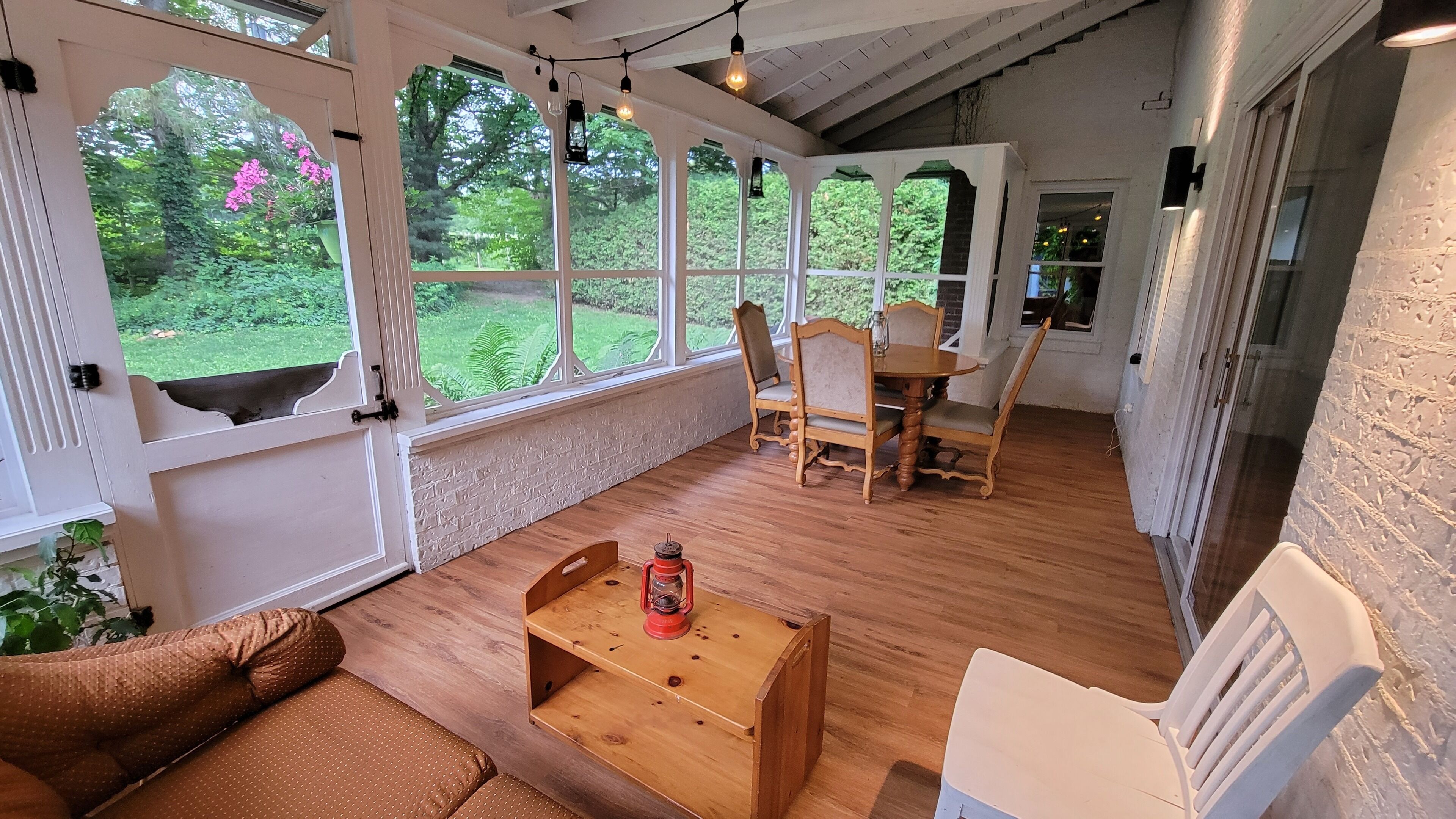 Ein gemütliches Zimmer mit Holzboden, einem Holztisch, einem Stuhl und einer Couch. Große Fenster bieten Blick ins Grüne.