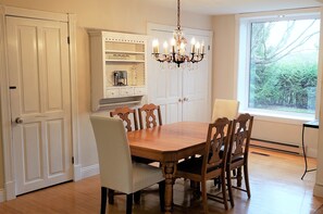A dining area with a wooden table and chairs, a chandelier, and a window with a view of trees. - Auberge du Vignoble Bromont (Brigham)