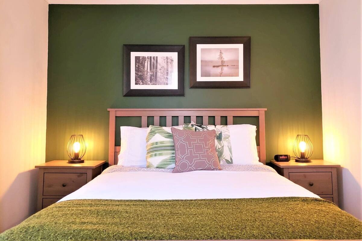 Un lit bien fait, recouvert d’une couverture verte, deux tableaux encadrés accrochés au mur et des lampes de chevet posées sur des tables de nuit en bois.