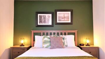 A neatly made bed with a green blanket, two framed pictures on the wall, and bedside lamps on wooden nightstands.