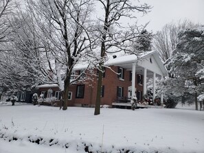 A brick house with a porch, surrounded by snow-covered trees. - Auberge du Vignoble Bromont (Brigham)
