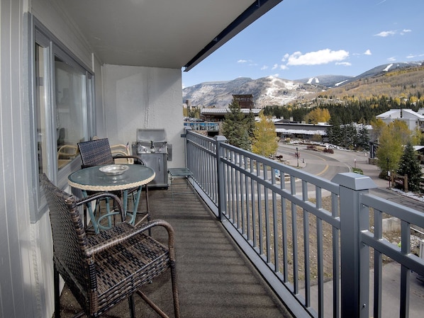 A balcony with wicker furniture, a grill, and a view of a town with mountains in the background.