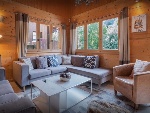 Interior acogedor de una cabaña con un sofá seccional gris, una mesa de centro de vidrio y un sillón de cuero. Amplias ventanas ofrecen vistas a árboles y una terraza de madera al exterior.