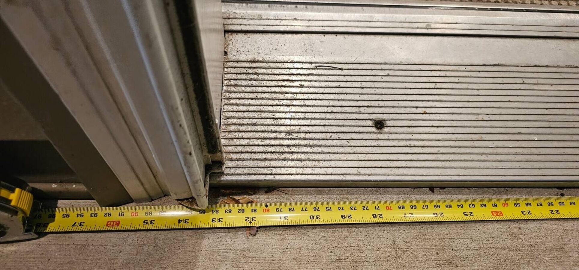 Uma fita métrica posicionada sob uma porta de metal, mostrando a largura da porta.