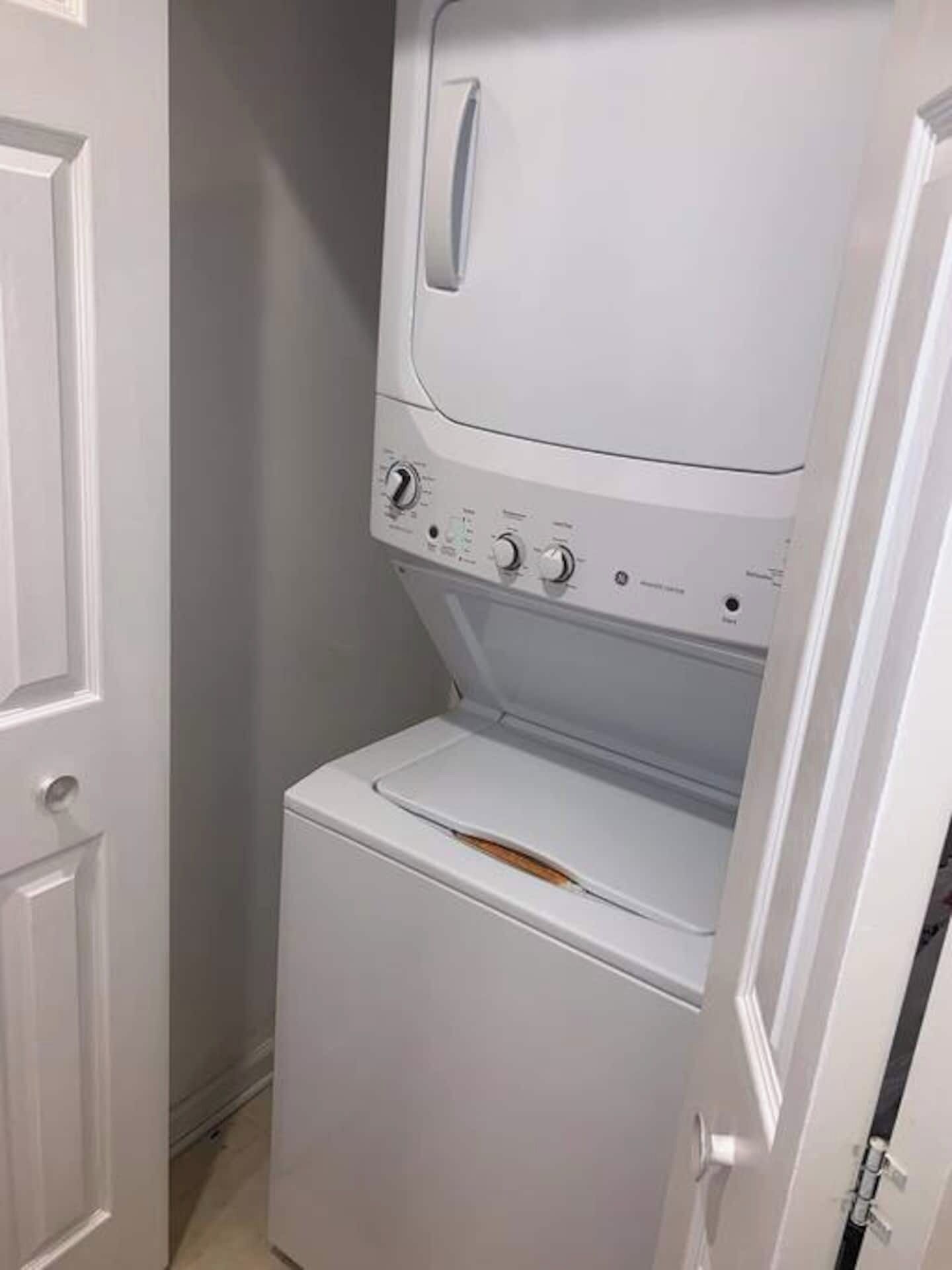 Een kleine, witte wasruimte met een voorlader wasmachine en droger, een bedieningspaneel met knoppen, en een deur met een zilveren handvat.