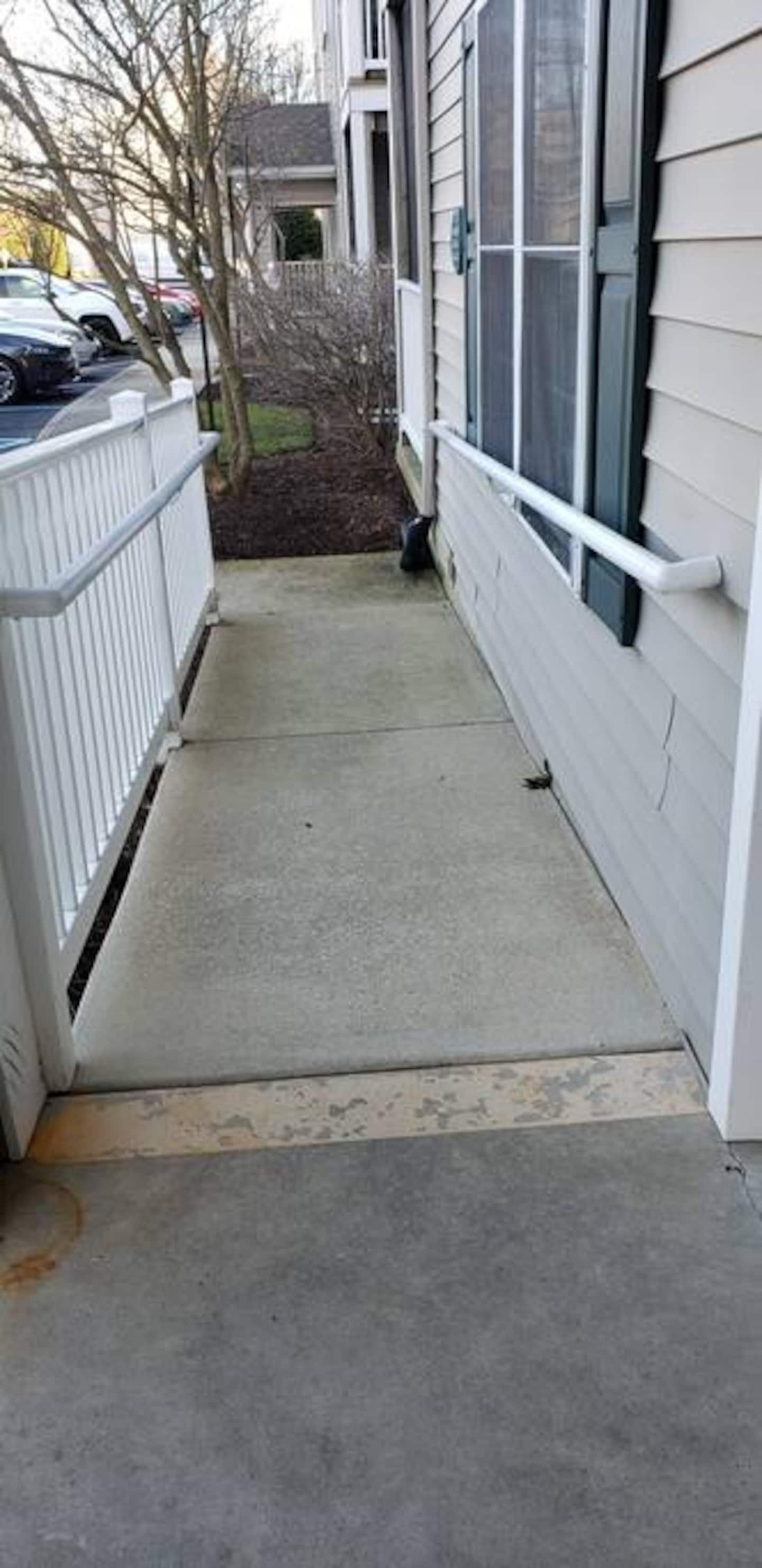 A narrow concrete pathway leading to a building with white siding and a white fence on one side.
