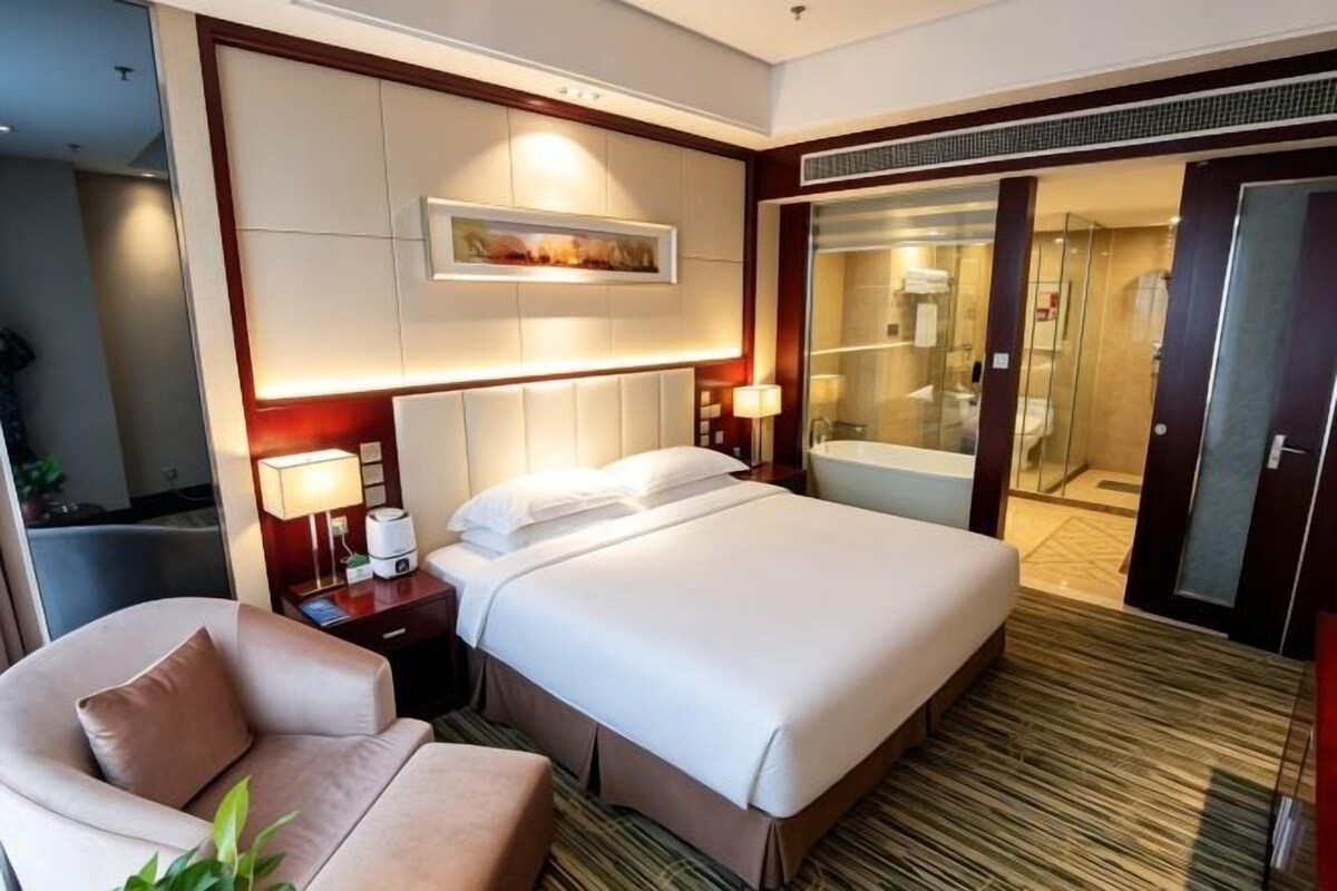 Habitación de hotel con una cama grande, un escritorio con lámpara, una silla, zona de ducha y una puerta de cristal que da al baño.