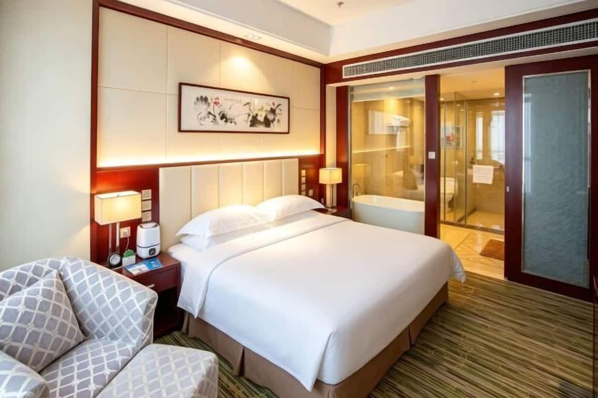 Habitación de hotel con una cama grande, un escritorio con silla, un baño con puerta de cristal y un cuadro en la pared.