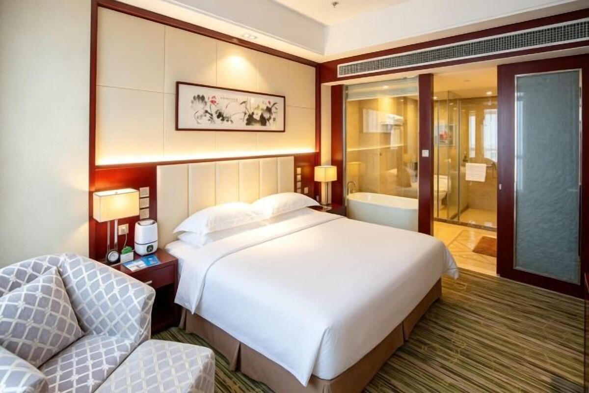 Une chambre d’hôtel avec un grand lit, un bureau avec une chaise, une salle de bain avec une porte en verre et un tableau encadré au mur.