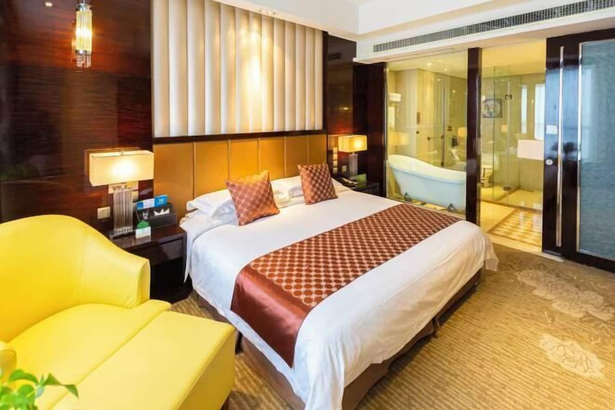 Habitación de hotel con una cama grande, un sillón amarillo, un baño con paredes de vidrio y una pared de acento de madera.