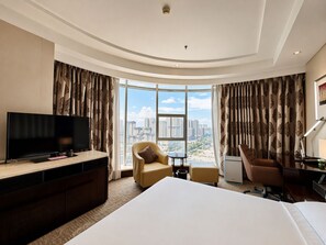 Une chambre d’hôtel avec un grand lit, une télévision, un bureau et une chaise. Les fenêtres offrent une vue sur le paysage urbain.