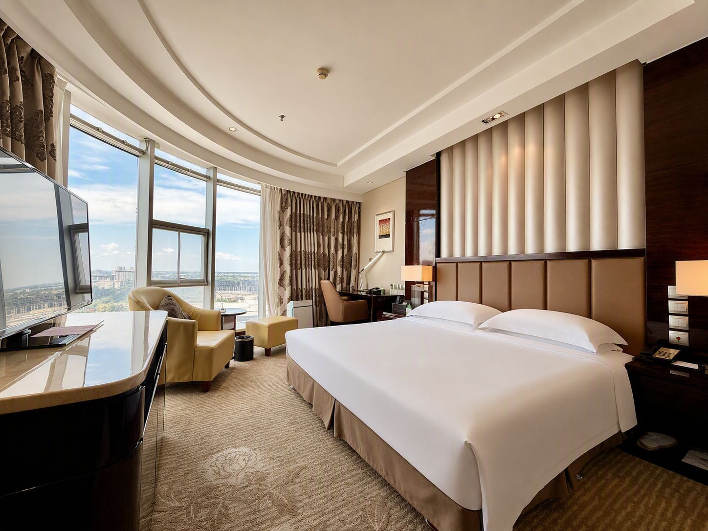 A hotel room with a large bed, a desk with a computer, a chair, a lamp, and a view of the city.