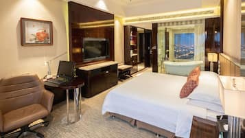 A hotel room with a bed, desk, chair, television, and a view of the city.