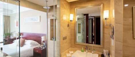 Habitación de hotel con cabina de ducha de vidrio, cama con ropa blanca, espejo grande y lavamanos blanco con artículos de aseo.
