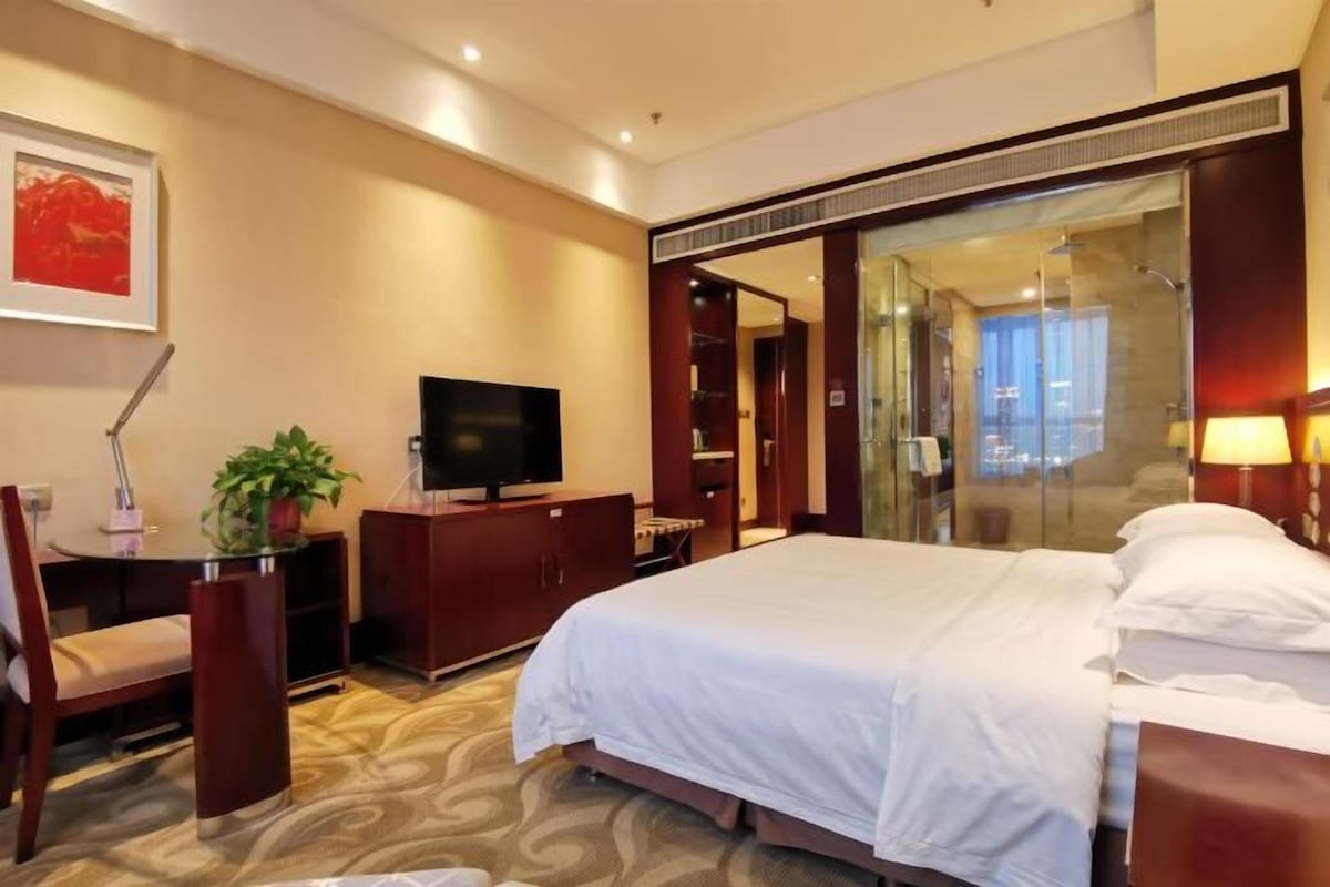 Une chambre d’hôtel avec un lit, un bureau avec une télévision, une chaise, une plante en pot et un grand miroir.
