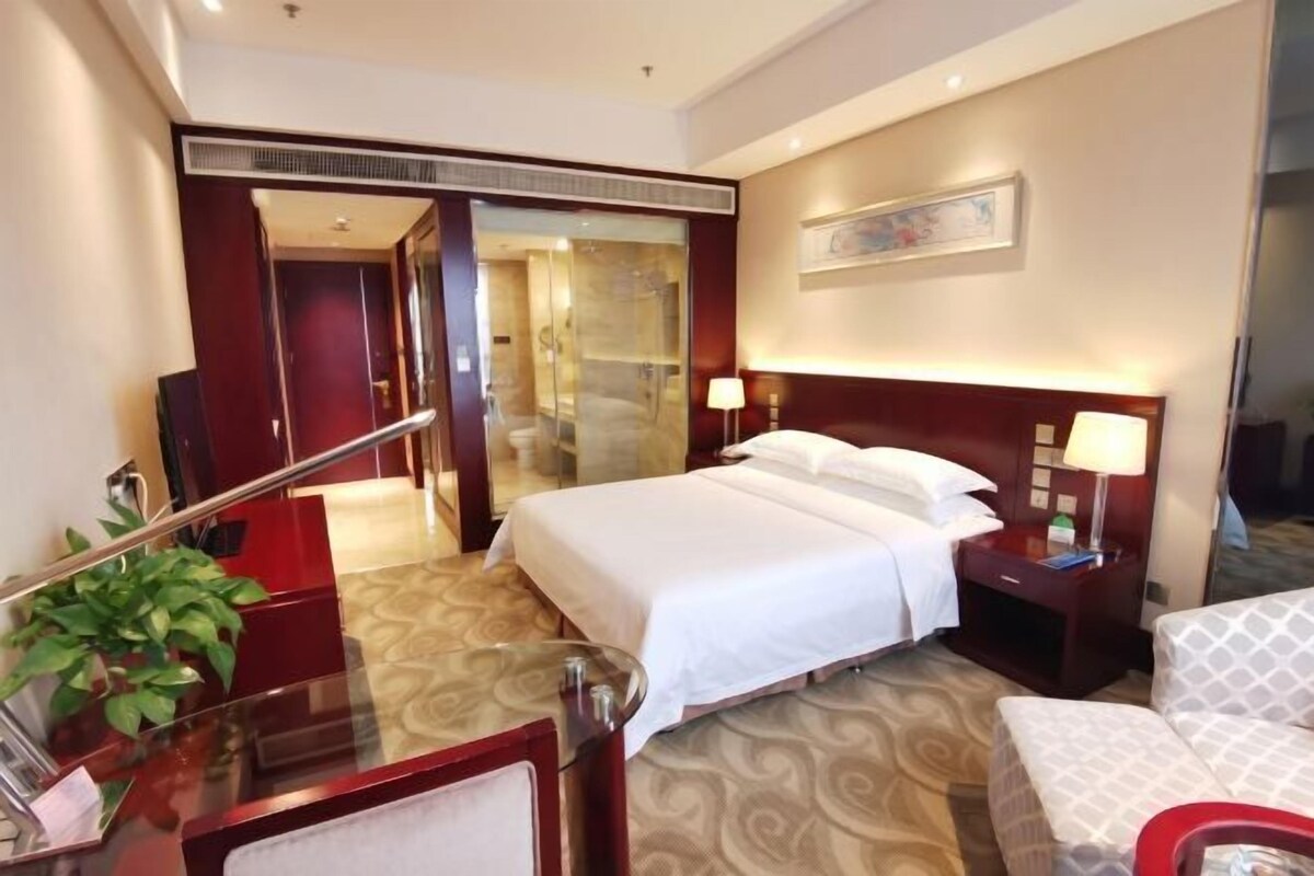 Habitación de hotel con cama, mesita de noche, lámpara y vista a un pasillo.