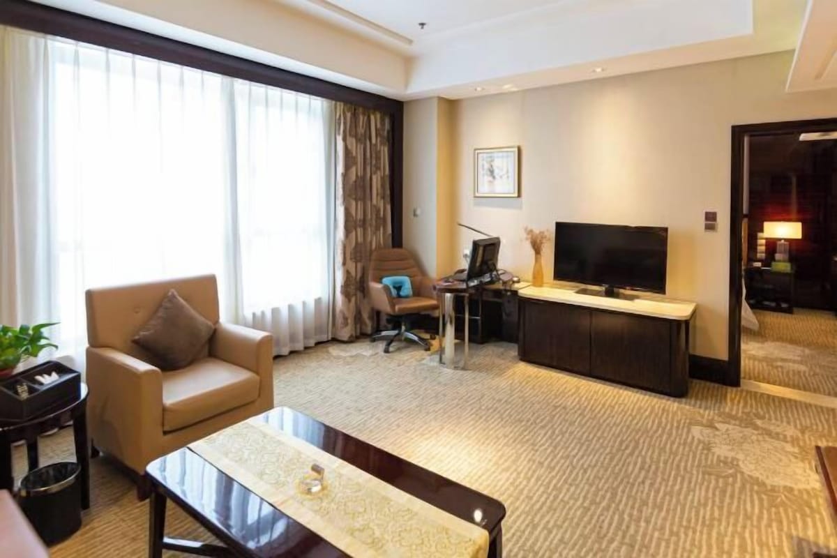 Une chambre d’hôtel équipée d’un canapé, d’une table basse, d’une télévision, d’un bureau avec un ordinateur et d’une grande fenêtre avec des rideaux.