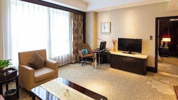 A hotel room with a sofa, a coffee table, a TV, a desk with a computer, and a large window with curtains.