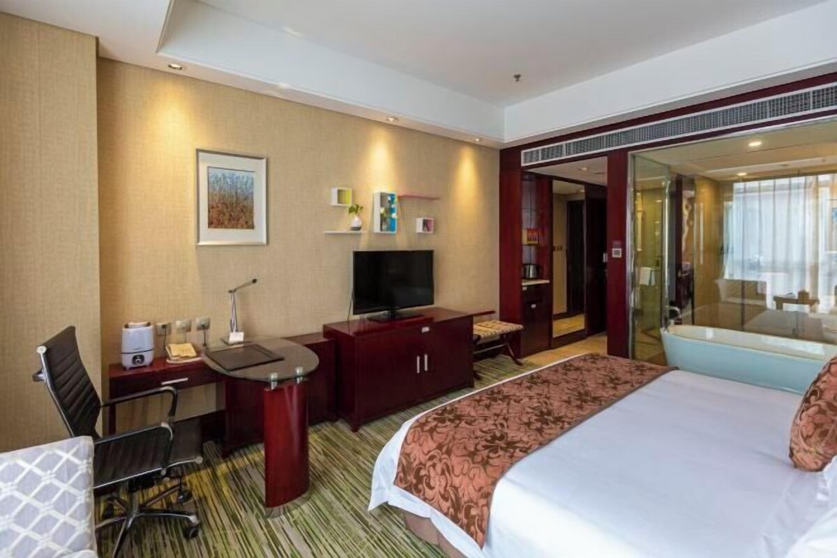 Habitación de hotel con cama, escritorio, silla, televisor y baño con puerta de vidrio.