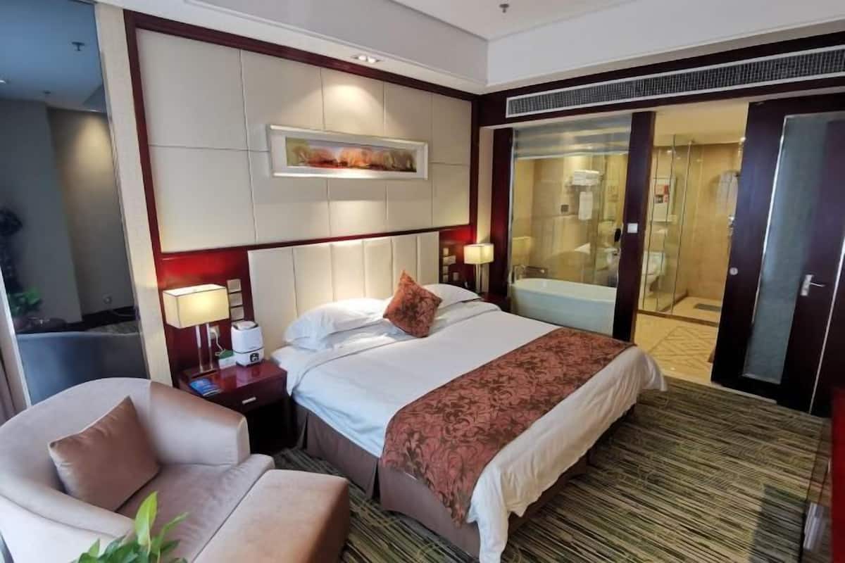 Una habitación de hotel con una cama grande, un área de descanso con una silla, un escritorio con una lámpara y un baño visible a través de una puerta de cristal.