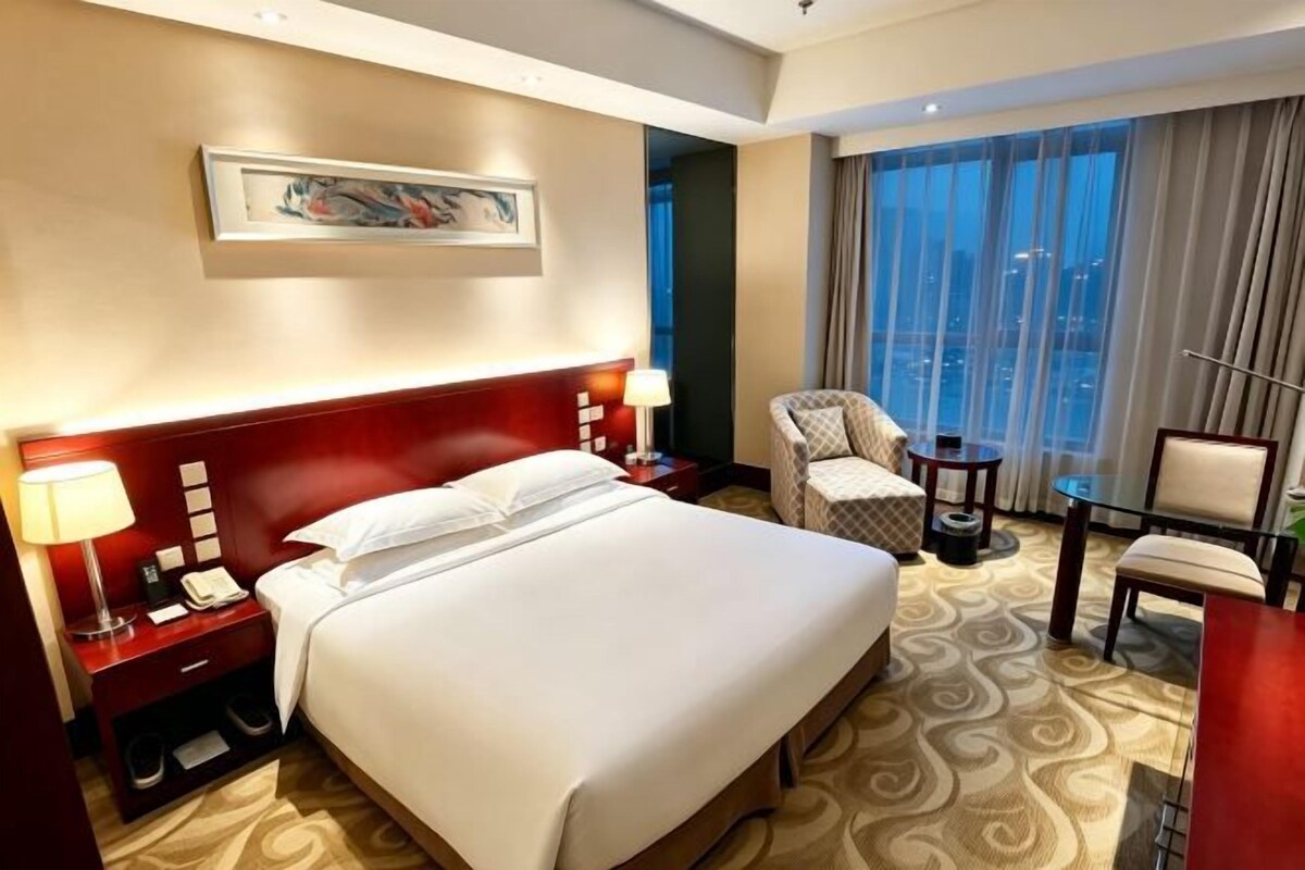 Une chambre d’hôtel avec un grand lit, des tables de chevet, une chaise, un bureau et une grande fenêtre avec des rideaux.