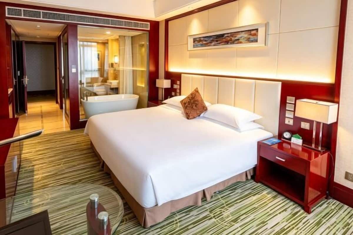 Habitación de hotel con una cama grande, una mesita de noche con lámpara, un baño visible a través de una puerta abierta y un cuadro en la pared.