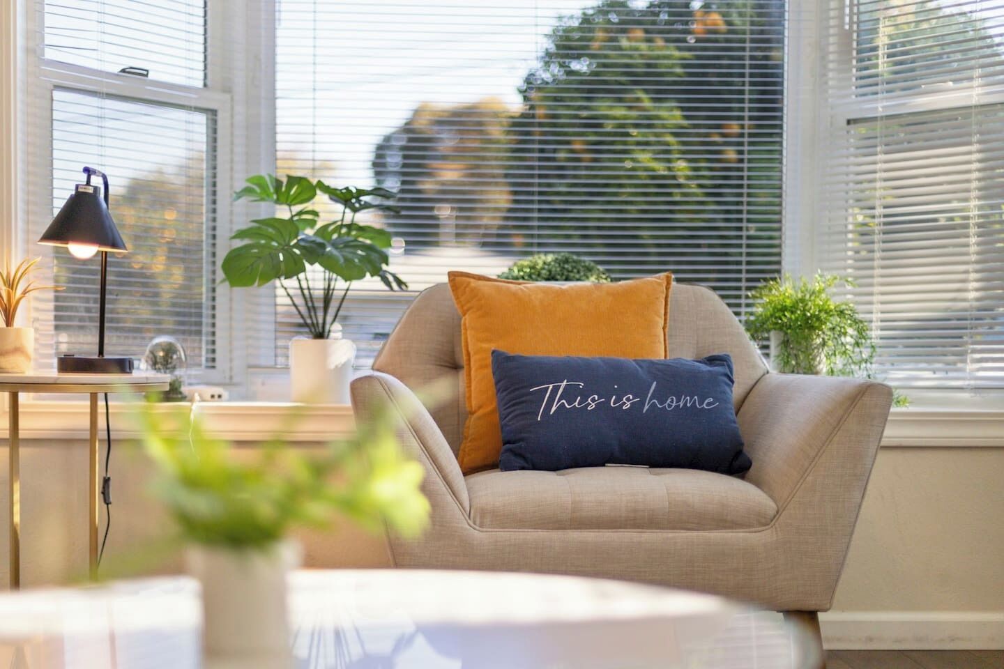 Ein gemütliches Wohnzimmer mit einem beigen Sofa, einem blauen Kissen mit dem Aufdruck "This is home" und einem kleinen Tisch mit einer Lampe und einer Pflanze.
