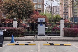 Estação de carregamento público com cabo conectado a um veículo, cercada por uma grade e vegetação.
