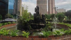A landscaped area with a rock formation and a fountain, surrounded by greenery and modern buildings in the background. - Jinhong Hotel (Zheng'an)