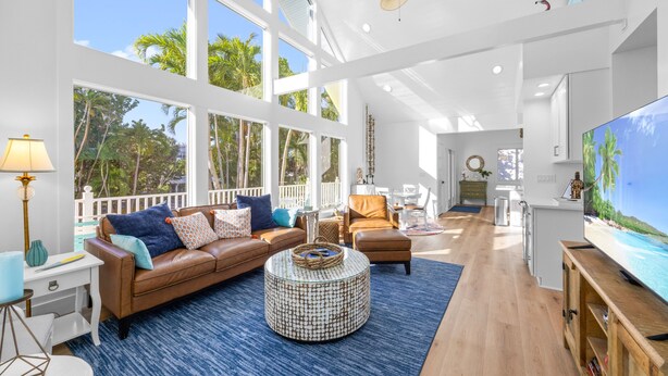 A spacious living room with a large TV, comfortable seating, and large windows offering a view of palm trees.