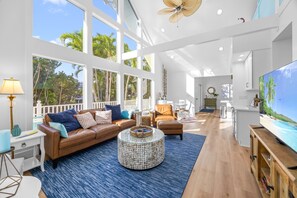 A spacious living room with a large TV, comfortable seating, and large windows offering a view of palm trees. - Six houses from stunning sandy Captiva Beaches ! (Captiva)