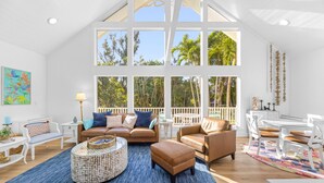 A spacious living room with a high ceiling, large windows, and a view of palm trees.