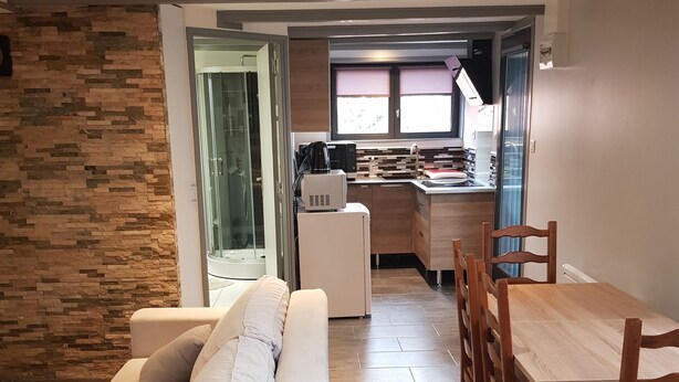 A modern living room with a sofa, dining table, and chairs. There is a kitchen area in the background with a window and a microwave. - Joli studio les brimbelles (Granges-Aumontzey)