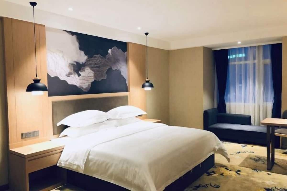 A hotel room with a bed, bedside table, lamp, artwork, and a view of the outdoors.