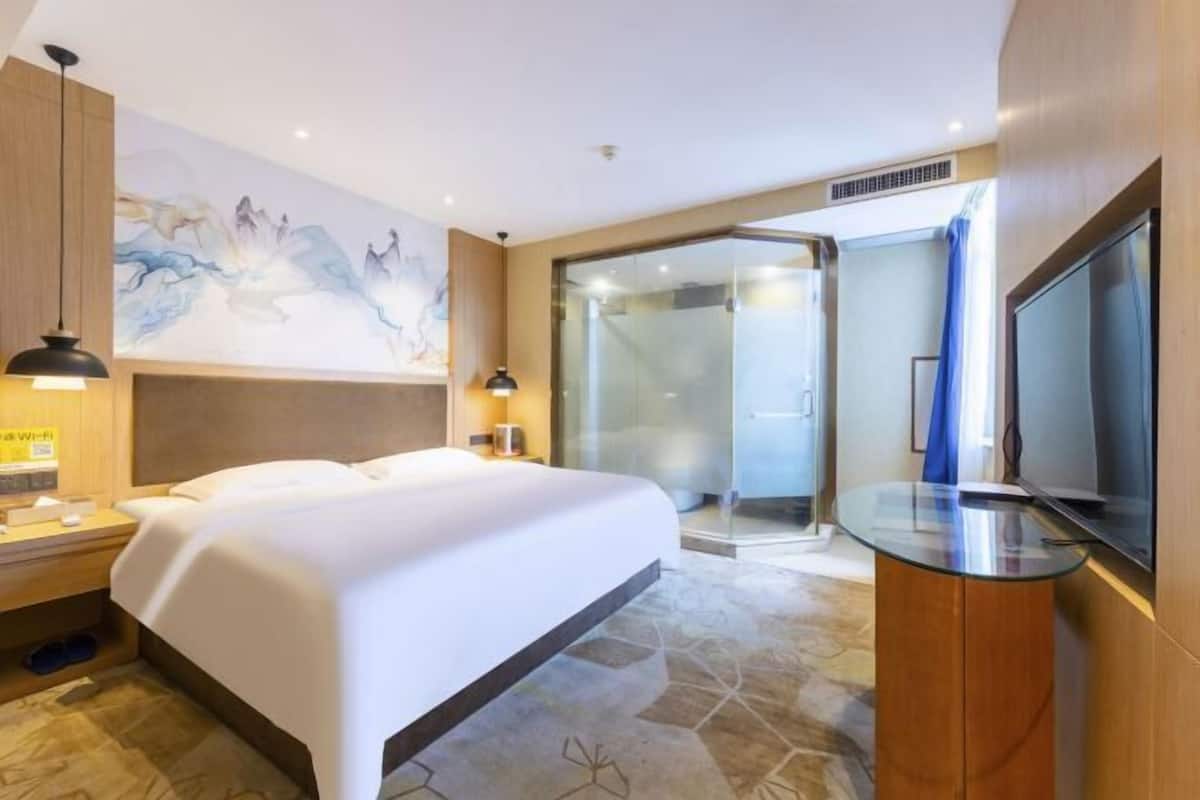 A hotel room with a bed, a TV, a glass shower enclosure, and a painting on the wall.