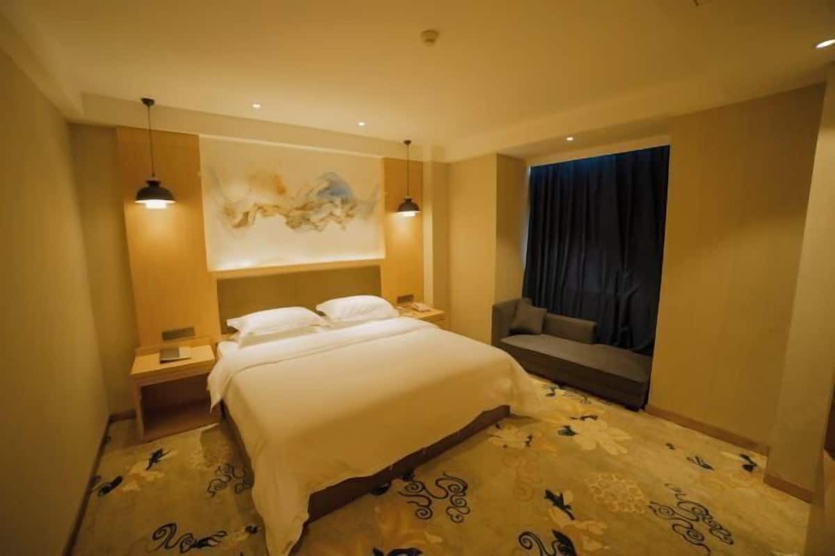 A hotel room with a large bed, a sofa, a wall-mounted artwork, and a window with curtains.