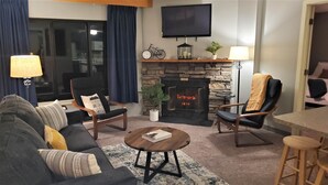 Una sala de estar con chimenea, un sofá, dos sillones, una mesa de centro y un televisor montado en la pared.