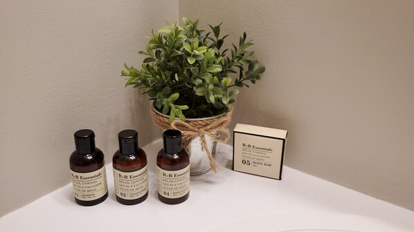 Trois flacons de produits de soins corporels de la gamme R&R Essentials, une plante en pot et une carte d’information sur le produit, le tout posé sur une surface blanche.