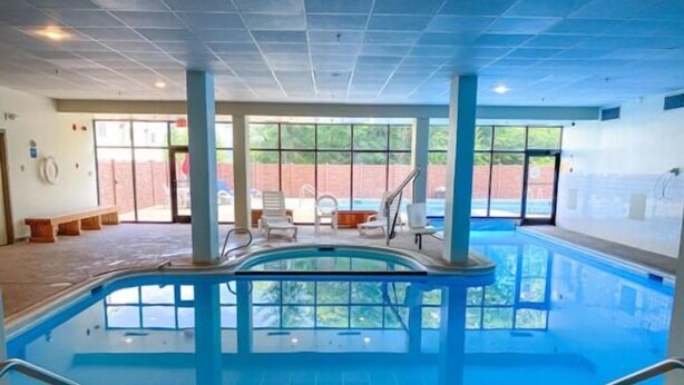 Una piscina interior con paredes de vidrio transparente, suelo de baldosas y algunas sillas de descanso.