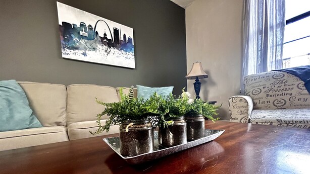 Ett vardagsrum med ett mörkt träbord som avlastningsbord, och på det står tre glasburkar med växter. Det finns en grå soffa med en blå kudde, en mönstrad fåtölj och en tavla med ett stadsmotiv på väggen.