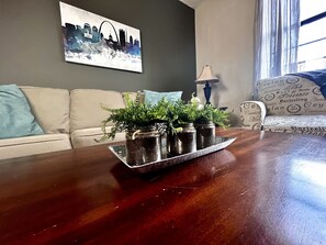 En stue med et mørkt træsofabord, der indeholder tre glas med planter. Der er en grå sofa med en blå pude, en mønstret lænestol og et bybillede på væggen.