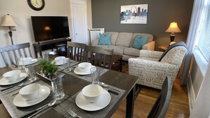 En matplats med ett bord dukat för fyra personer, en soffa, en tv och en tavla med en stadsbild på väggen.